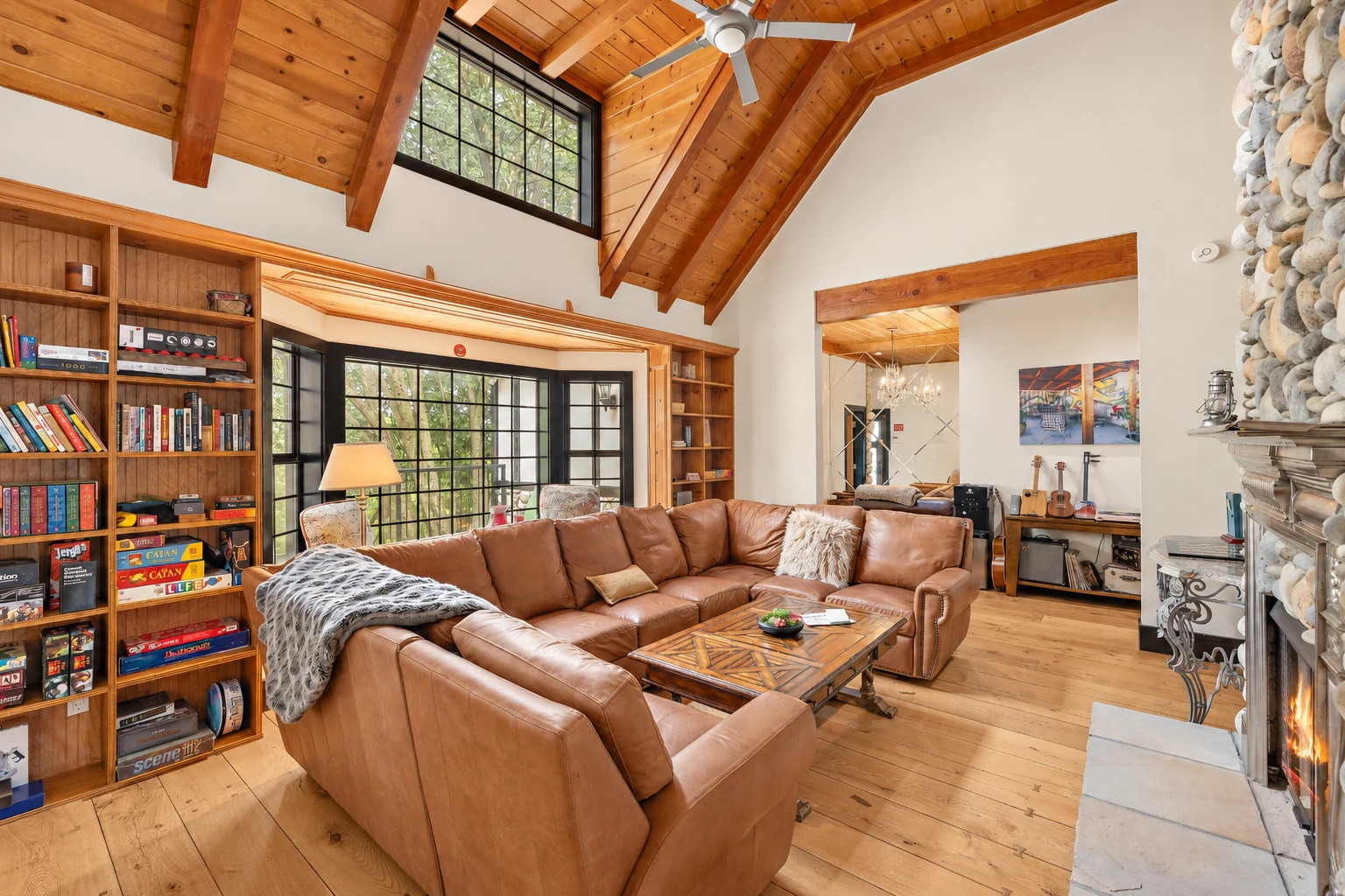 A living room with a vaulted wood ceiling, leather sofas, built-in bookshelves and a river-stone fireplace
