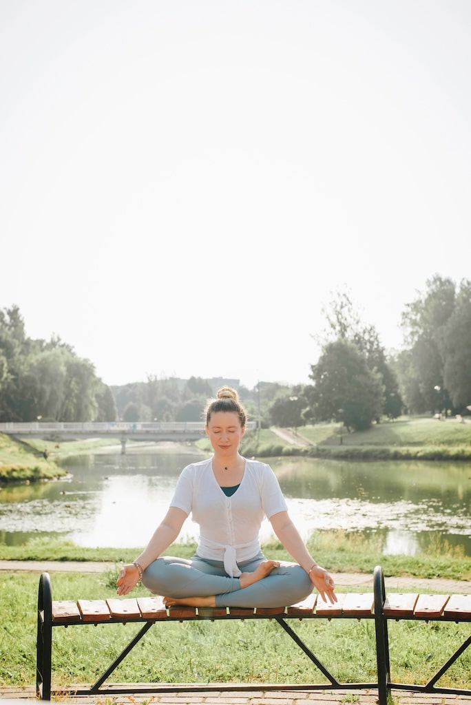 Happy young woman sitting on a bench in lotus position and meditating in the morning outdoors