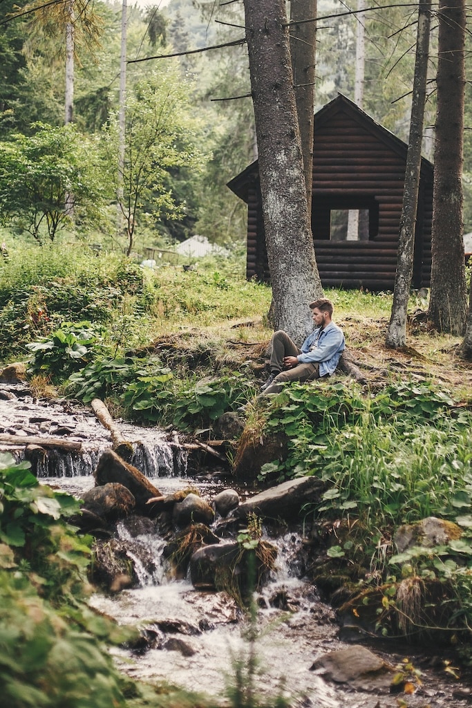 Man sitting at river in forest on background of wooden log in mountains. Exploring woods in summer. Copy space. Travel and wanderlust concept. Relaxing with nature