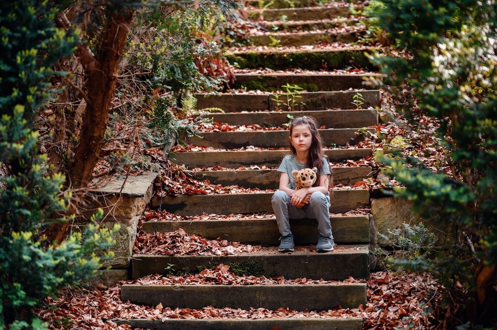 Young girl sitting on the steps outside with her teddy bear, signifying childhood trauma and sadness.