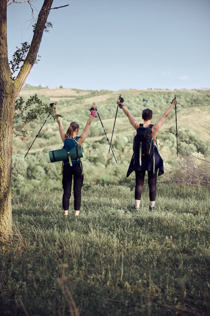 Rear view of a two female friends hikers with trekking poles celebrating with raised hands after getting on the top of the mountain.