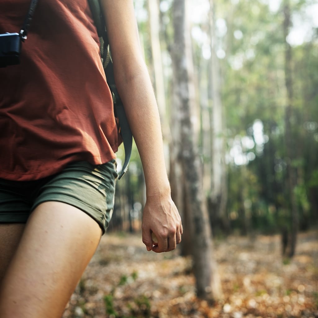 Individual walking through the woods surrounded by trees, isolated close up photo of hand, backpack, and leg walking.