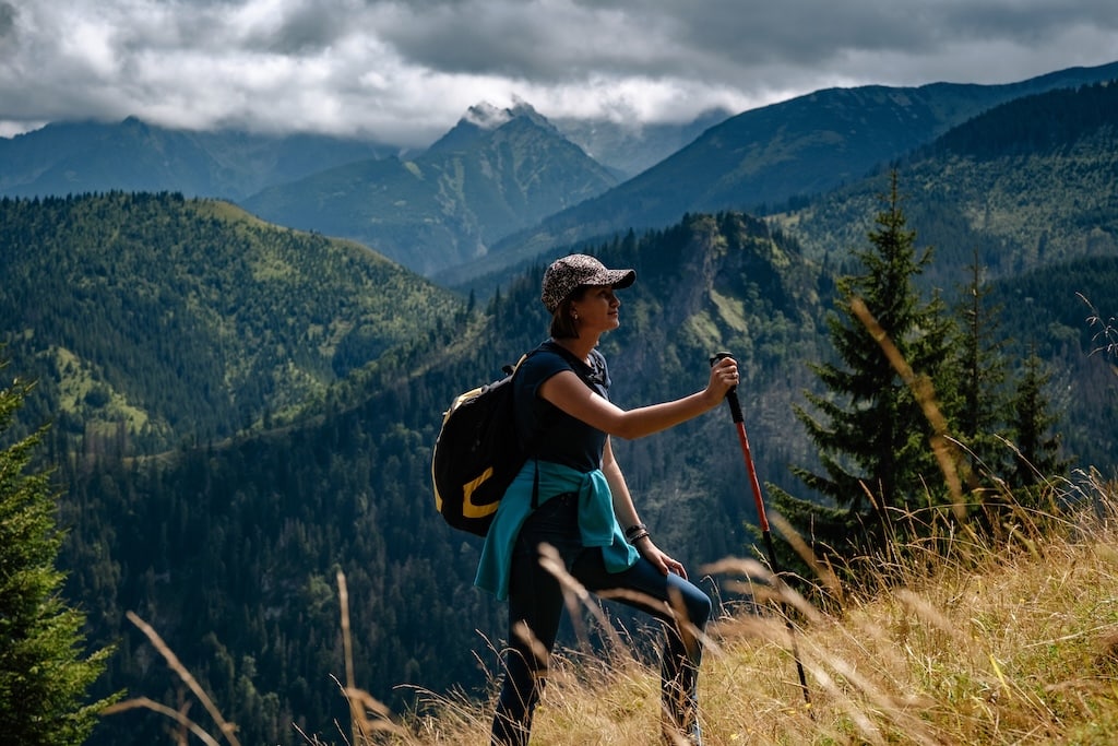 A young woman with a backpack and a tracking stick is climbing a mountain.