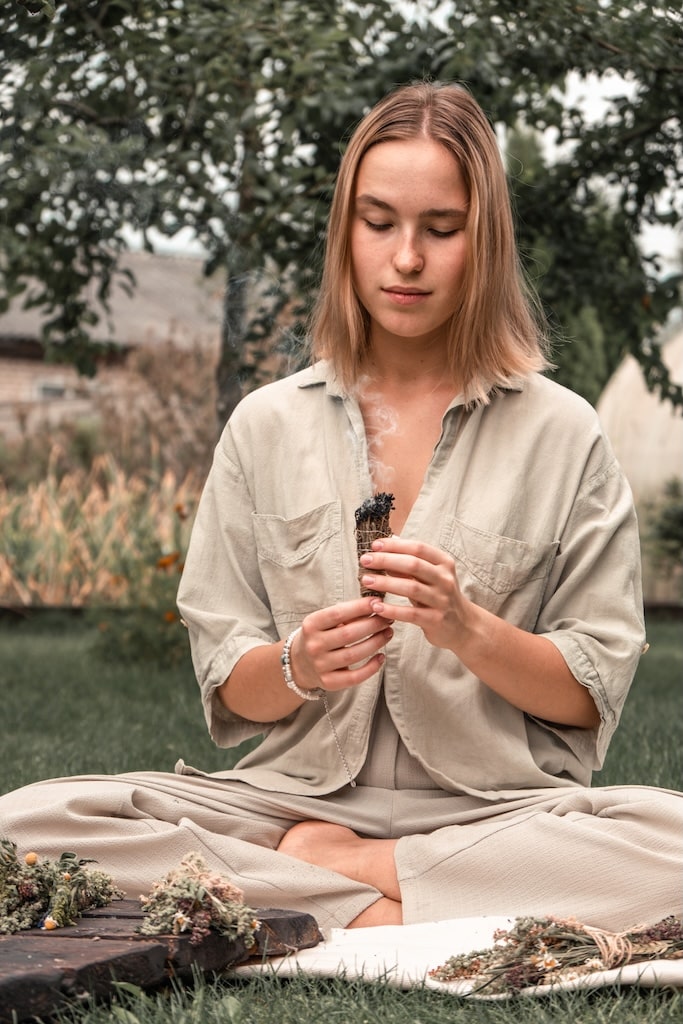 Woman Inhaling Incense Smoke During MeditationGround level of relaxed female meditating and breathing while sitting in Lotus pose near fragrant incense during yoga session in garden