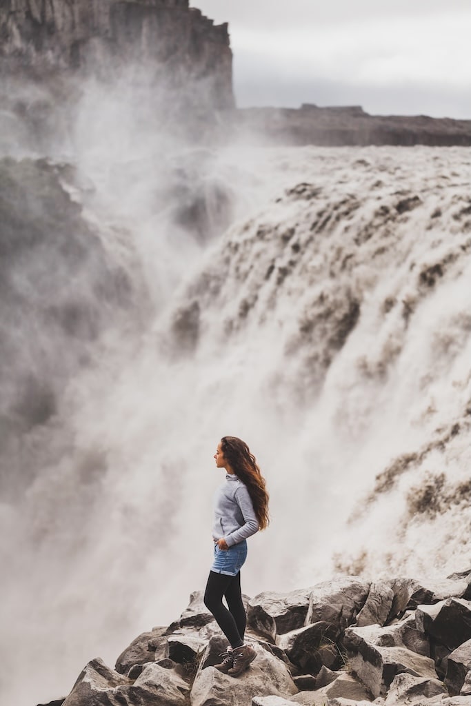 Young woman in jeans shorts and gray hoodie with dramatic view of waterfall